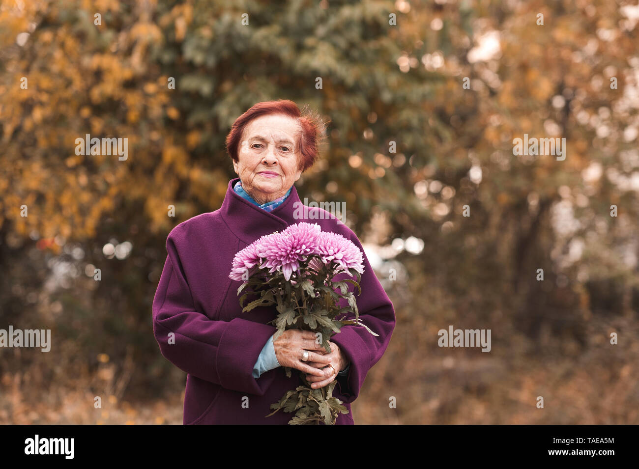 Sorridente donna senior 70-80 anni elegante da indossare giacca invernale azienda fiori all'esterno. Guardando alla fotocamera. Foto Stock