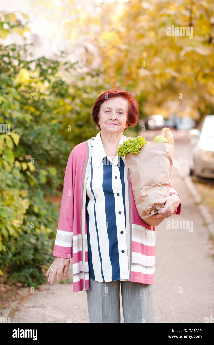 Senior donna 70-80 anno vecchia azienda sacchetto di carta con acquisti a casa a piedi all'esterno. Guardando alla fotocamera. La stagione autunnale. Foto Stock