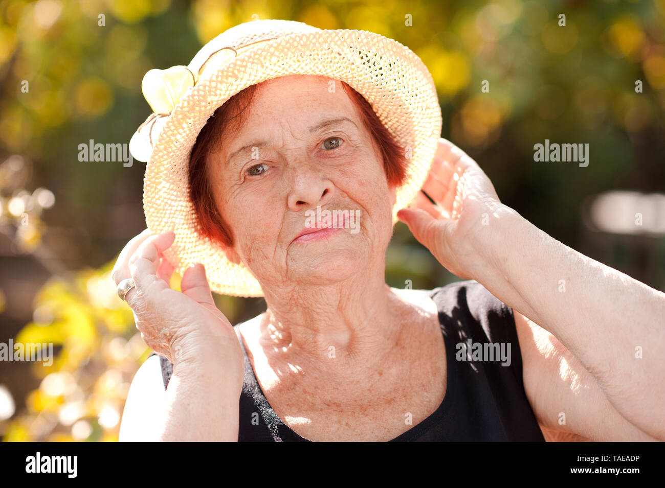 Estate ritratto di sorridere senior donna che indossa cappello di paglia su verde natura. Guardando alla fotocamera. In posa all'aperto. 70s. Foto Stock