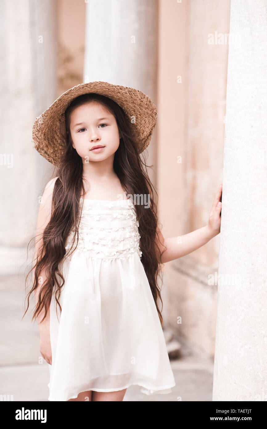 Carino kid girl 4-5 anno indossando cappello di paglia e di elegante abito bianco all'esterno. A piedi nella parte antica della città. Guardando alla fotocamera. Foto Stock