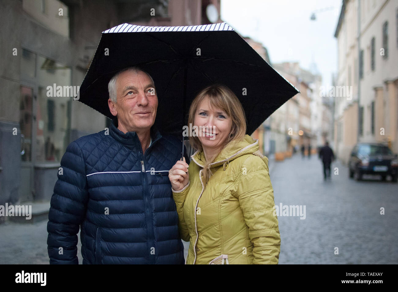 Donna di mezza età e anziani il suo marito di trascorrere del tempo insieme all'aperto in piedi sotto il loro ombrello su strada asfaltata. Coppia con differenza di età Foto Stock
