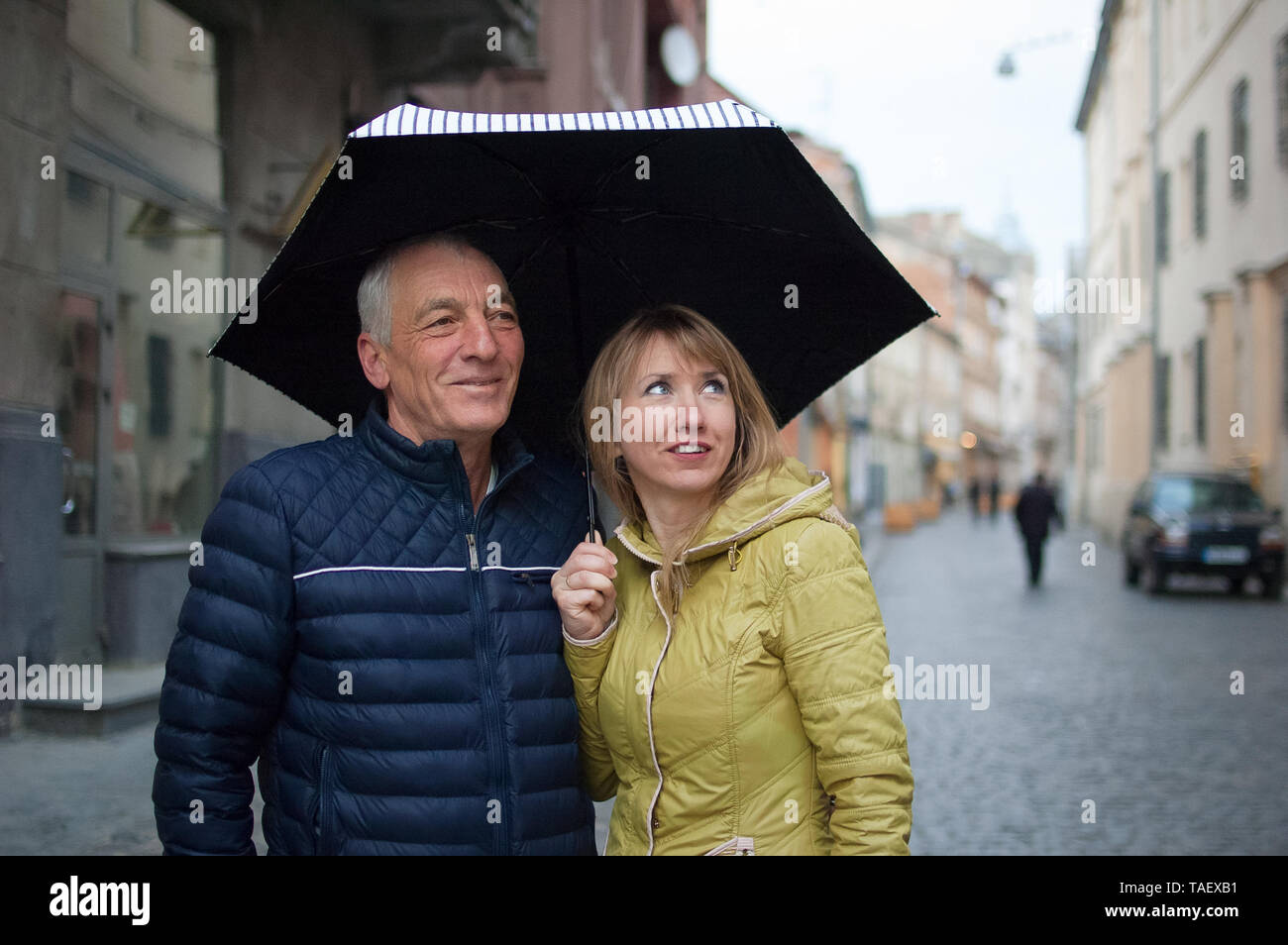 Close up ritratto di uomo anziano e il suo giovane biondo e moglie che abbraccia ogni altra e in piedi sotto il loro ombrello su strada asfaltata. Coppia con Foto Stock
