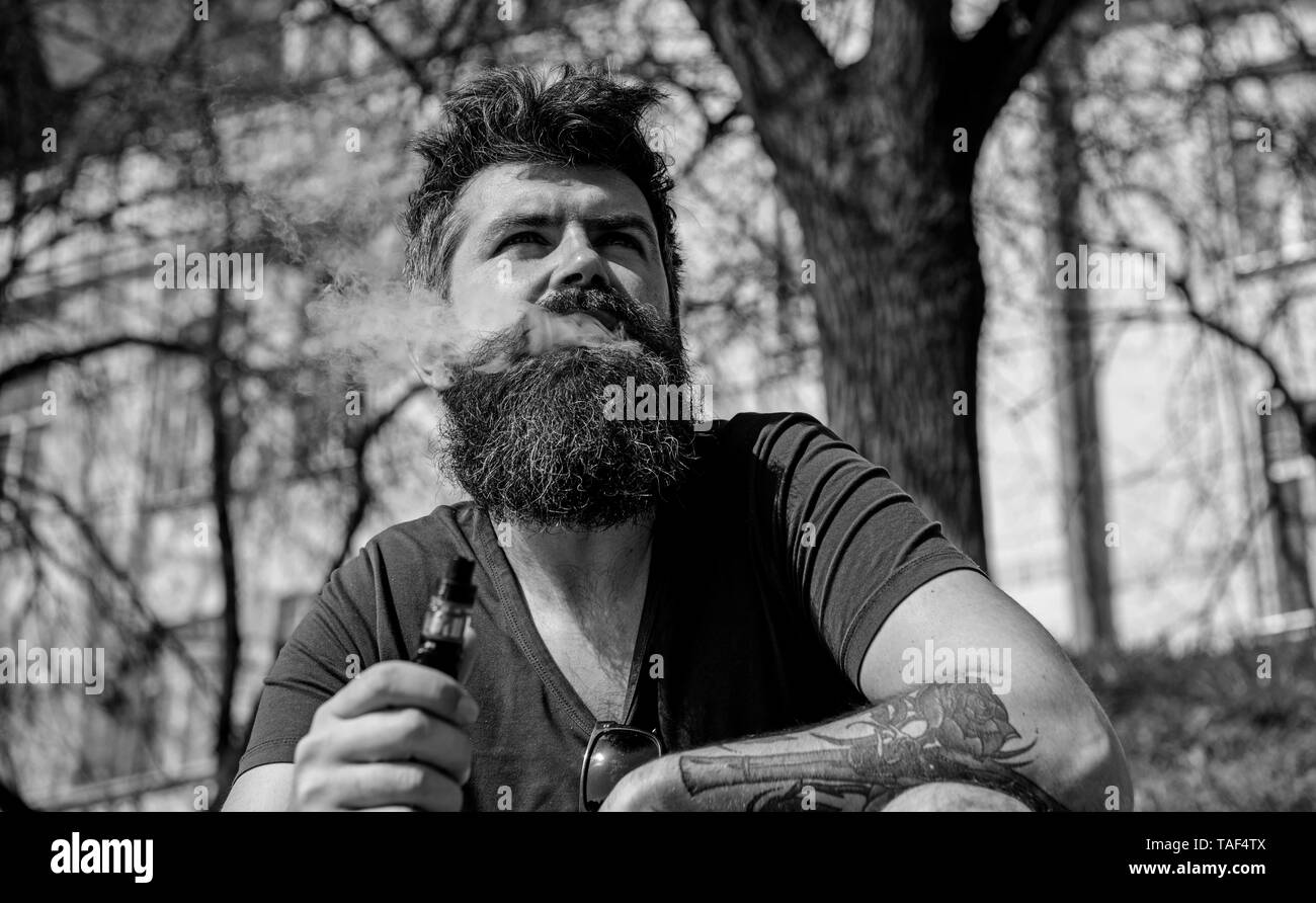 Hipster con tousled capelli e grigio su rilassato faccia sognante con emissione di fumo bianco volare al di fuori della bocca. Vaping e abitudini del concetto. Uomo con barba e baffi fumatori, background urbano. Foto Stock