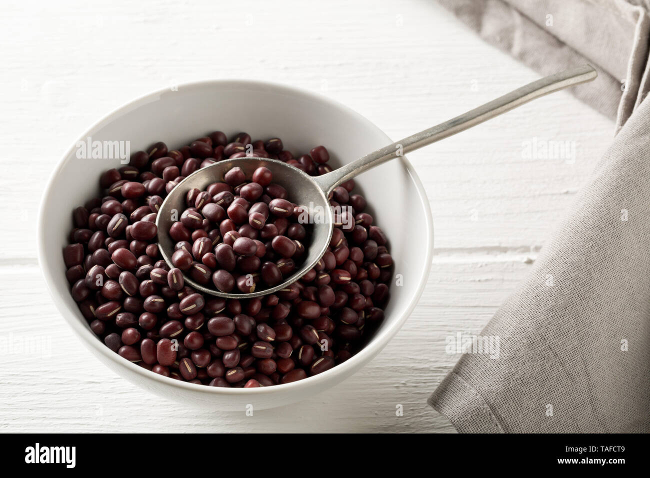 Materie, non cotte, essiccato adzuki (rosso) Mung bean in bianco ciotola con cucchiaio in metallo su legno bianco sullo sfondo della tabella con il fuoco selettivo Foto Stock