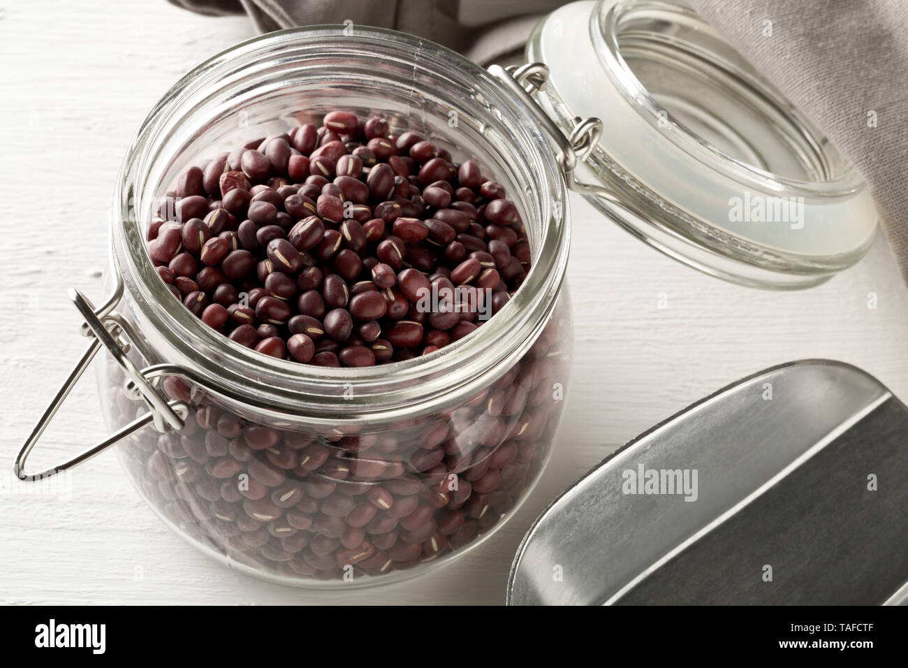 Materie, non cotte, essiccato adzuki (rosso) Mung bean in vetro la memorizzazione di vaso con cucchiaio in metallo bianco sul tavolo in legno con sfondo grigio asciugamano da cucina Foto Stock