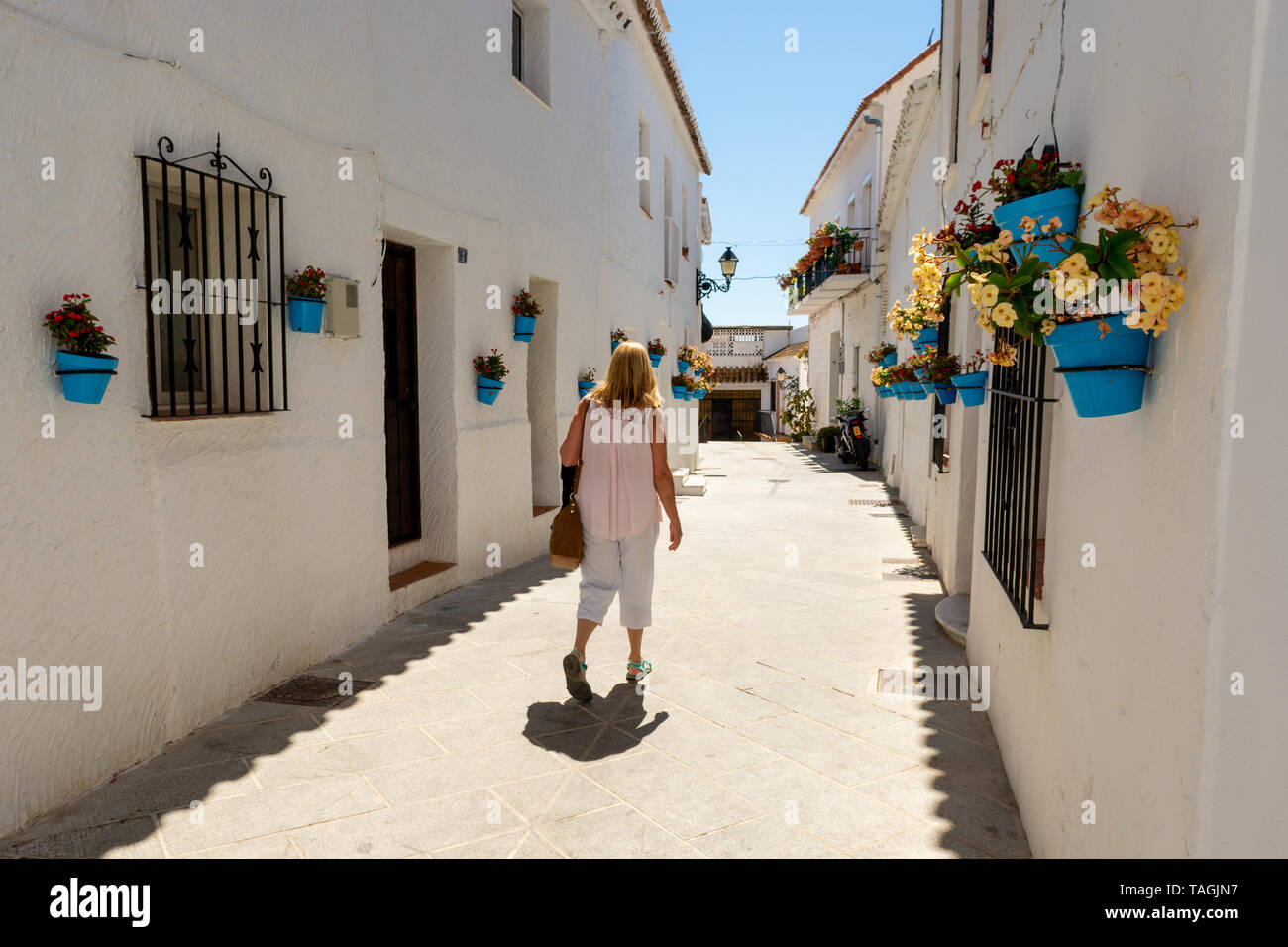 Donna che cammina giù per una strada stretta con impianto di blu vasi appesi sulle pareti bianche nel pittoresco villaggio di montagna di Mijas, regione Andalusia, Spagna Foto Stock