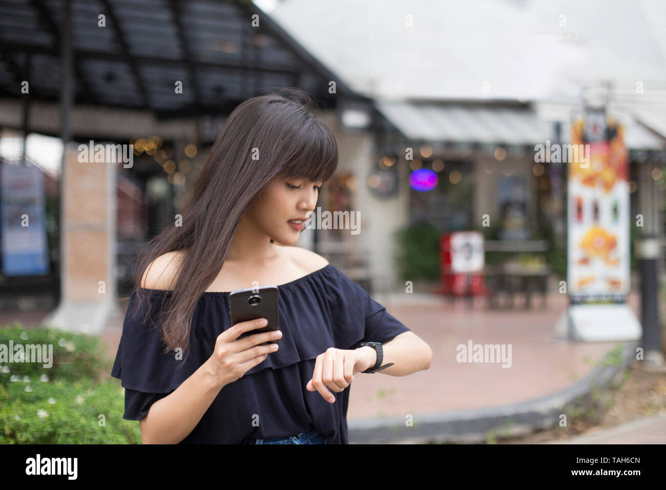 Giovane bella donna asiatica tenendo lo smartphone e guardando un orologio da polso, in attesa di qualcuno Foto Stock