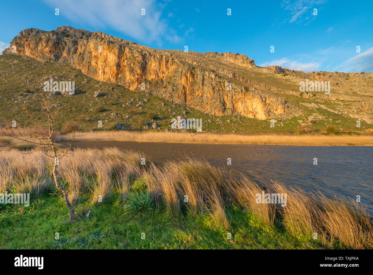 Tramonto in Grecia, ora d'oro in montagna, piccolo lago con erba bruciata, vento forte. Picco di montagna con parete dorata rock. Foto Stock