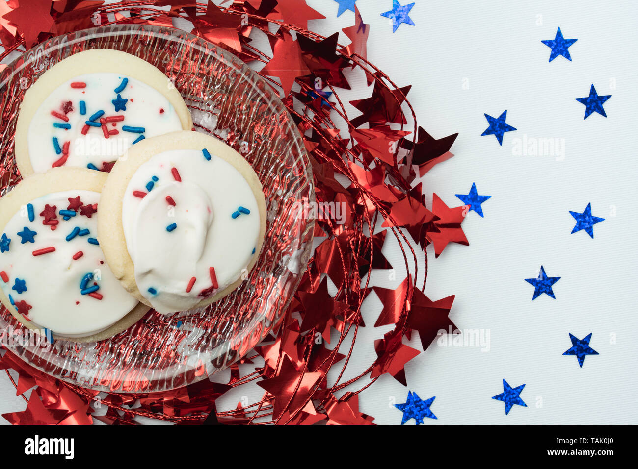 Brillante lucido stella rossa garland sotto una lastra di vetro con zucchero biscotti ricoperti di glassa bianca e spruzza e blu glitter stelle su un tavolo bianco Foto Stock