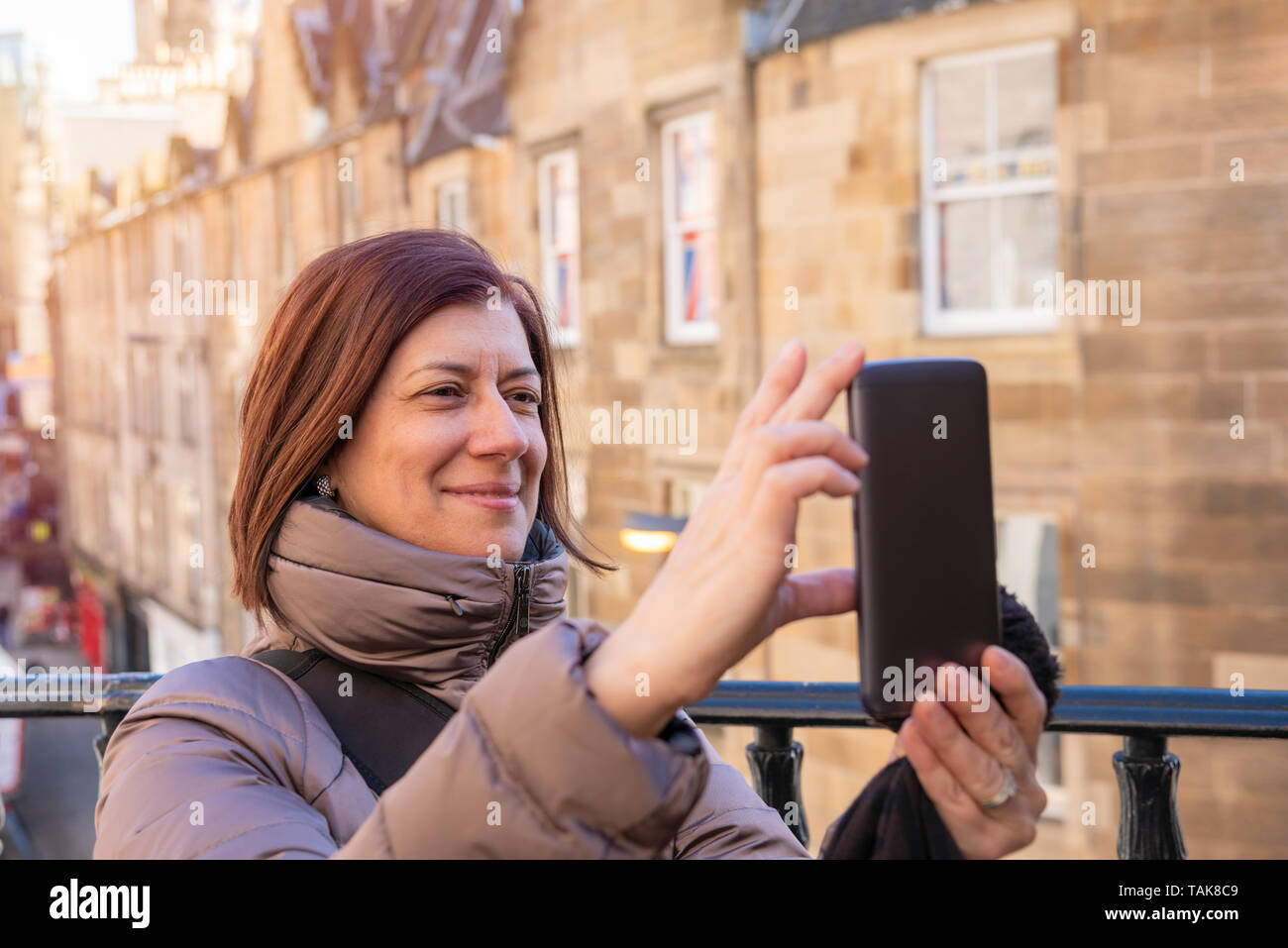 Donna sorridente turistica prendendo un selfie in un centro storico con buildins in background su una soleggiata giornata invernale Foto Stock