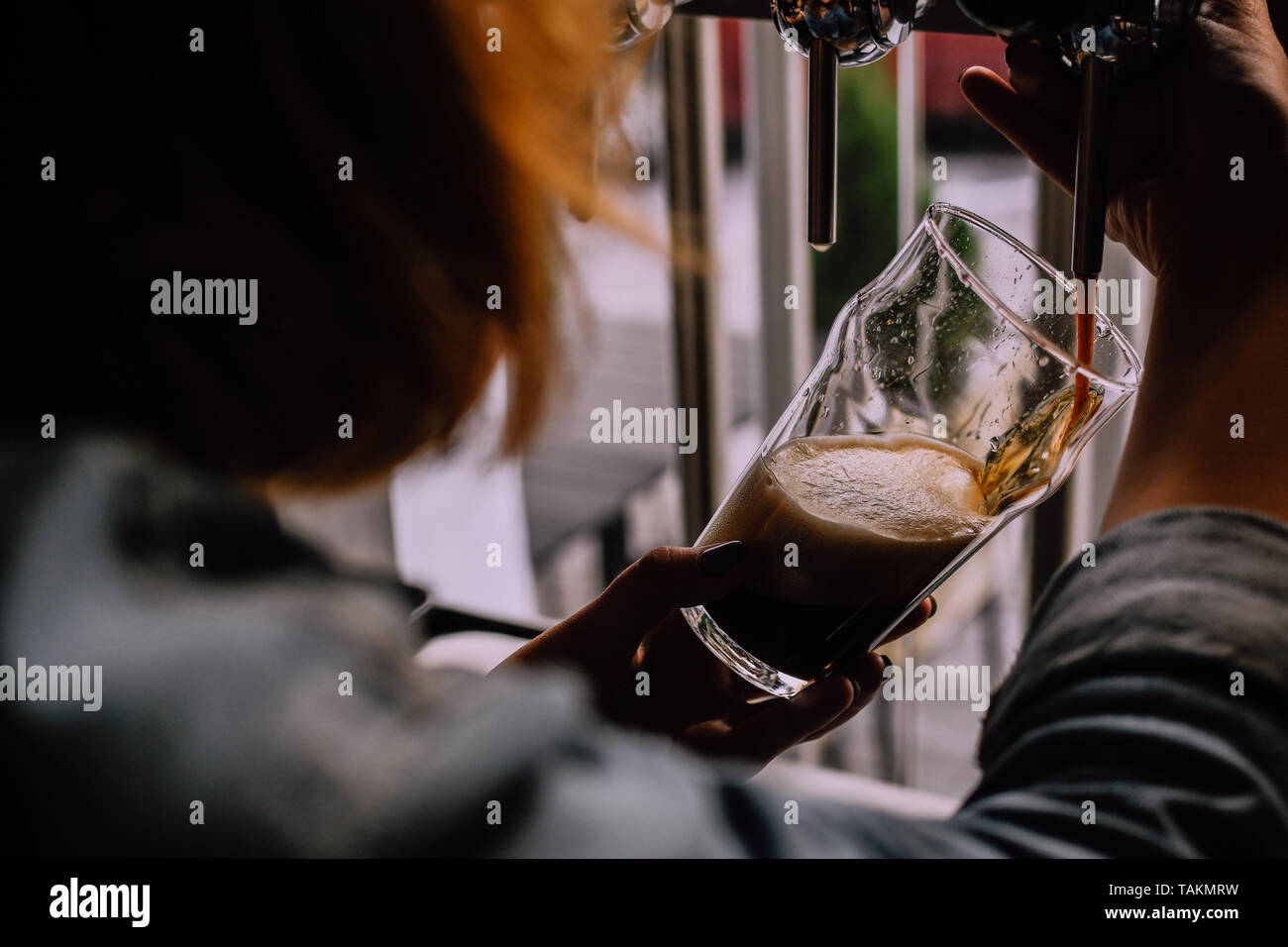 Ragazza di colare una pinta di birra scura in una birra artigianale pub. Pub locale. Lucido rubinetti birra sullo sfondo. Luminosa e soleggiata e pomeriggio. Guarda da una spalla Foto Stock