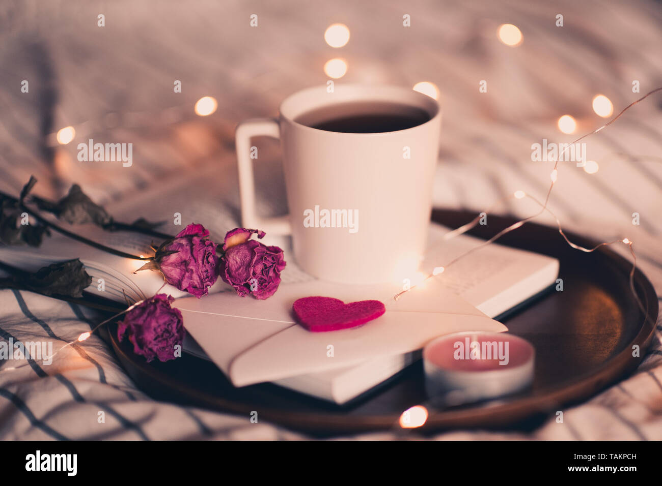 Tazza di tè che soggiornano sul libro aperto con i fiori e la busta in letto oltre le luci sul background. Il giorno di San Valentino. Foto Stock