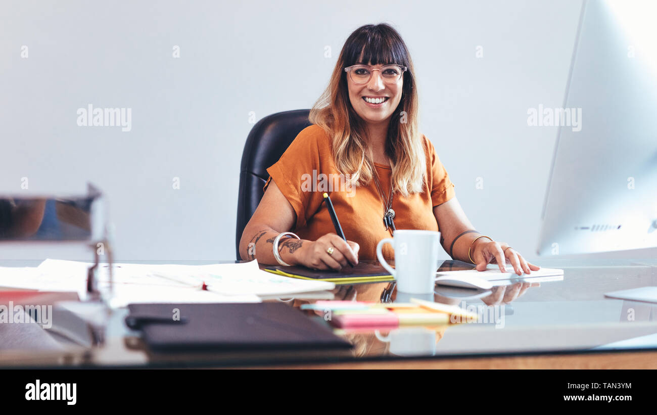 Illustrator fare uno schizzo su un blocco di scrittura digitale. Donna sorridente imprenditore lavorando su i suoi disegni seduto alla sua scrivania in ufficio. Foto Stock