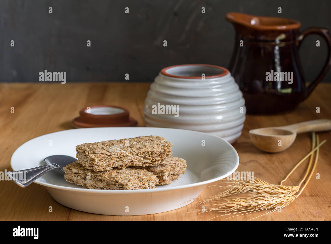 Frumento barrette di cereali a secco in piastra colazione sul tavolo di legno con spighe di grano in primo piano e il miele e latte jar in background Foto Stock