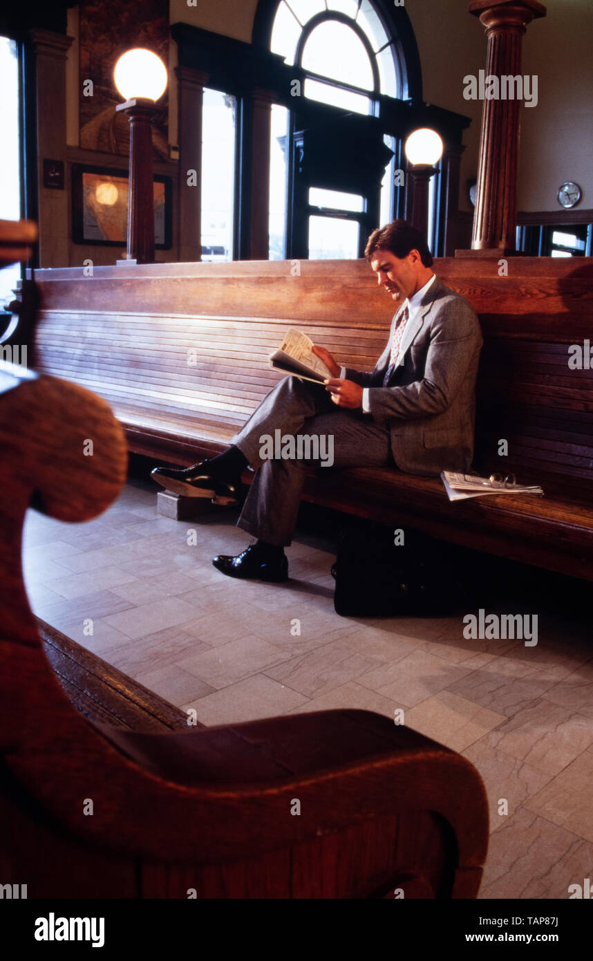 Imprenditore in abito grigio legge giornale finanziario in attesa per il treno in stazione terminale in sala d'attesa. Foto Stock