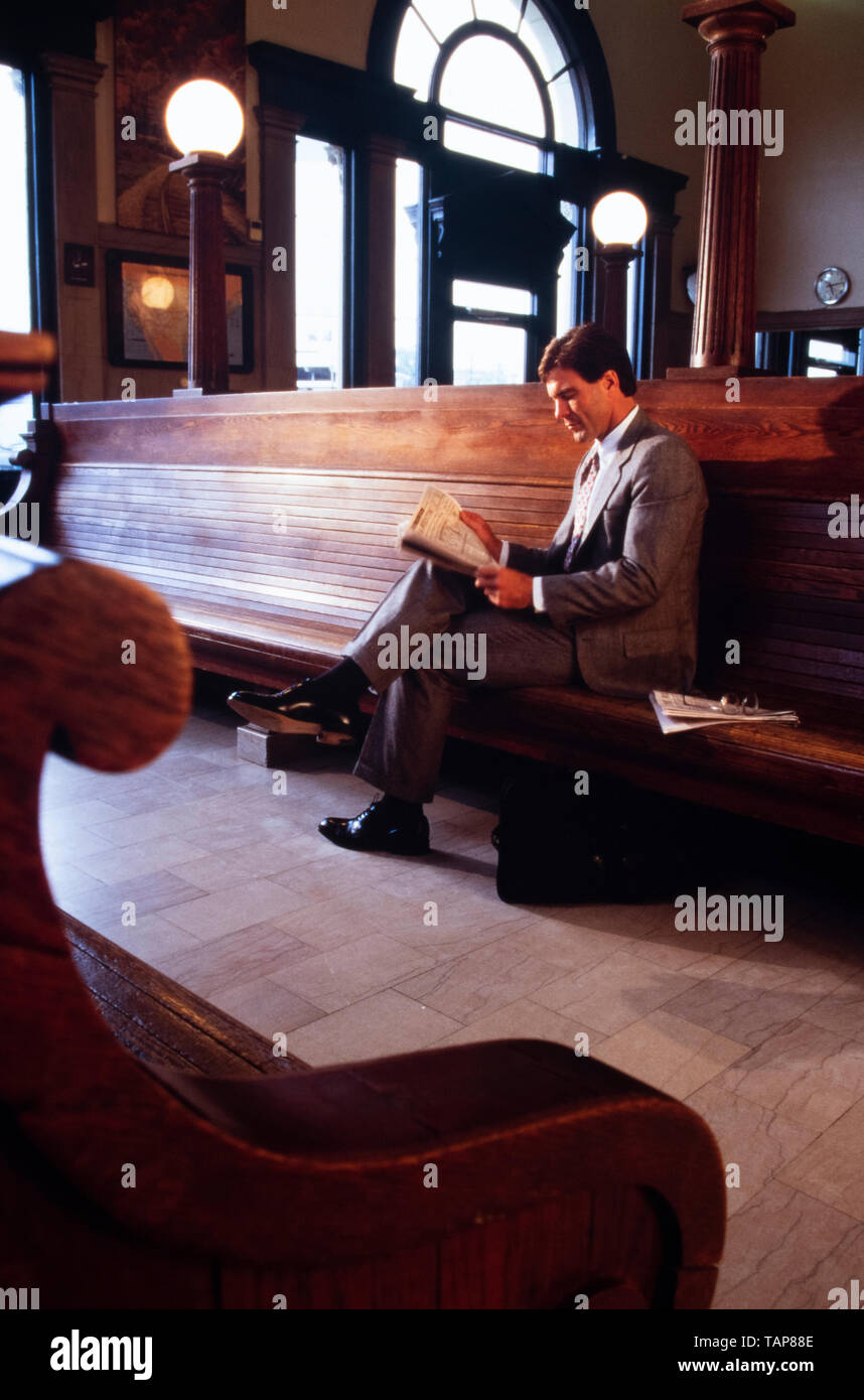 Imprenditore in abito grigio legge giornale finanziario in attesa per il treno in stazione terminale in sala d'attesa. Foto Stock