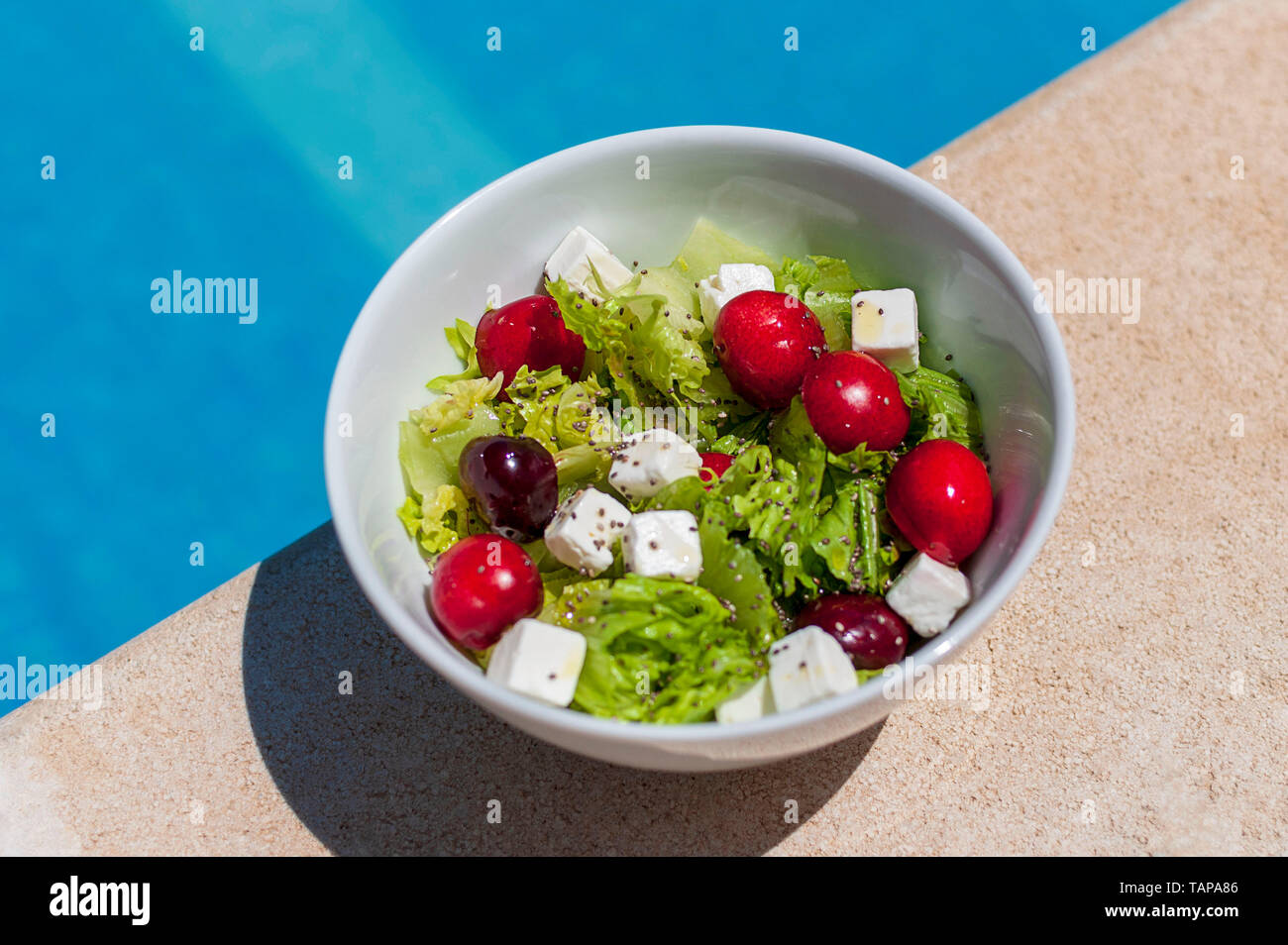 Fresca e sana insalata in un isolato ciotola bianco. A lato della piscina, in una giornata di sole. La lattuga, semi, Big Red e succose ciliegie, feta c Foto Stock