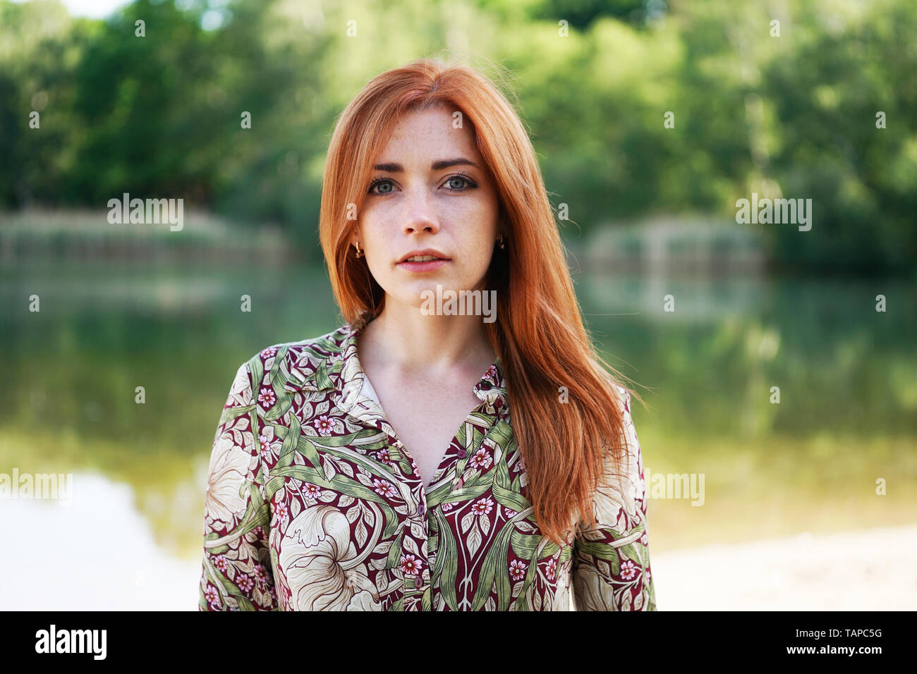 Raffreddare e fiducioso giovane donna indossa motivo floreale abiti estivi con lunghi capelli rossi in piedi dal lago - autentico persone reali Foto Stock