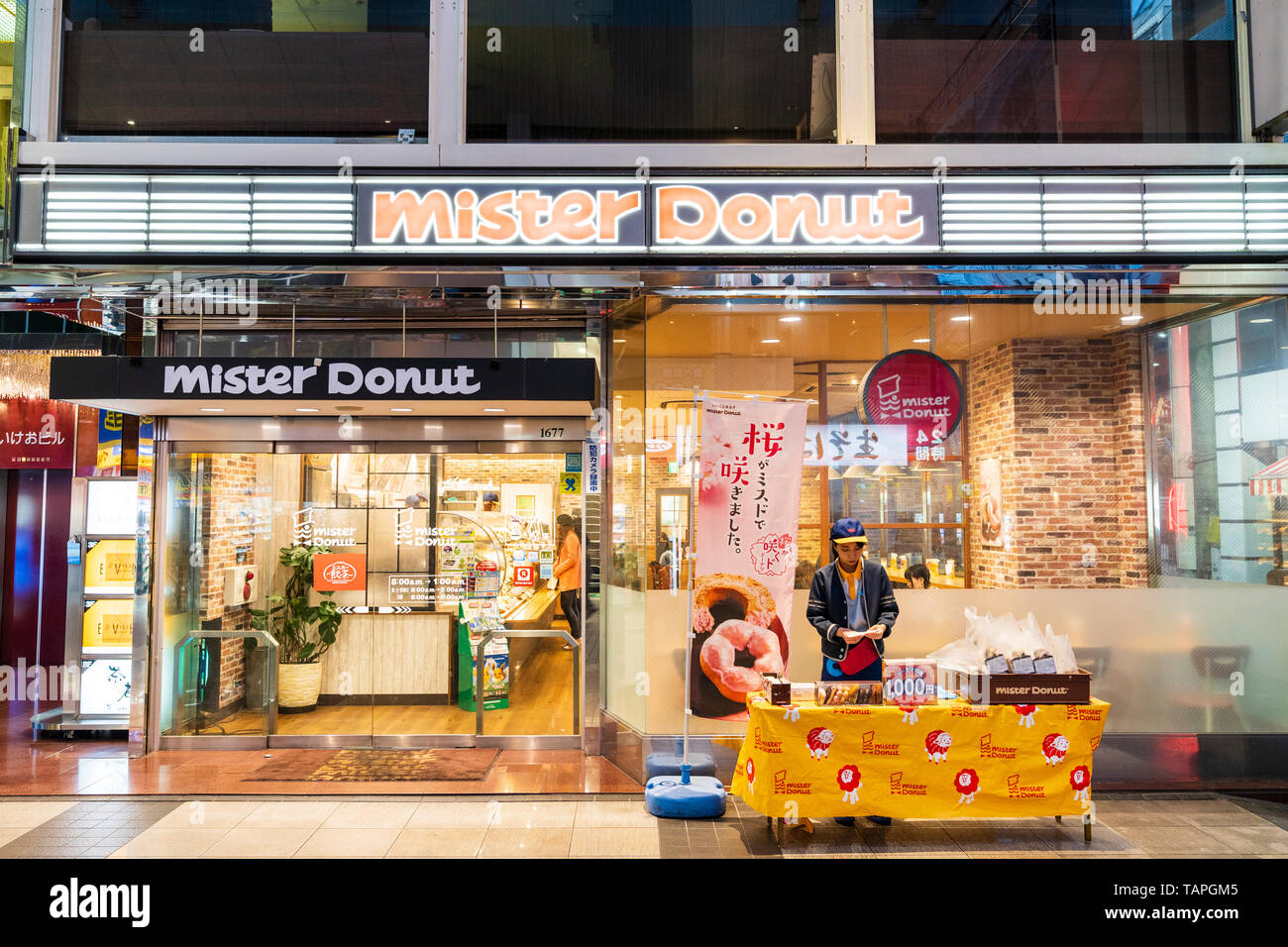 Kumamoto, Giappone. Illuminata di notte, un open Mister Donut diner con piccoli take away stand al di fuori del ristorante. Le persone all'interno di sedersi e mangiare. Foto Stock