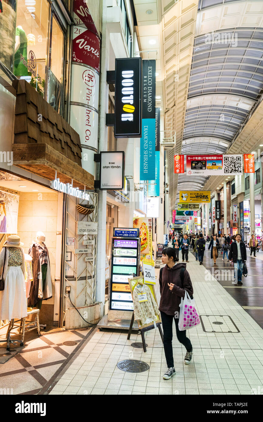 Vista lungo un lato del Shimotori galleria con negozi e boutique alla moda in primo piano. Arcade occupato con la gente, di notte. Kumamoto, Giappone. Foto Stock
