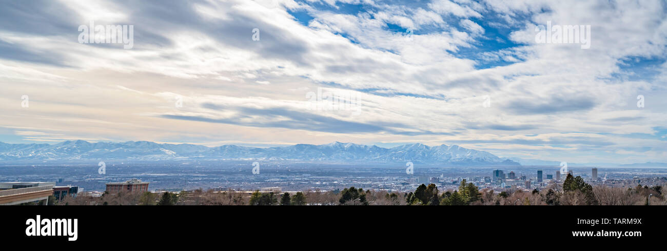 Vista panoramica di una vivace città incorniciata da lussureggianti alberi in primo piano. Un lontano Snow capped mountain può anche essere visto sotto la nuvola riempì il blu Foto Stock