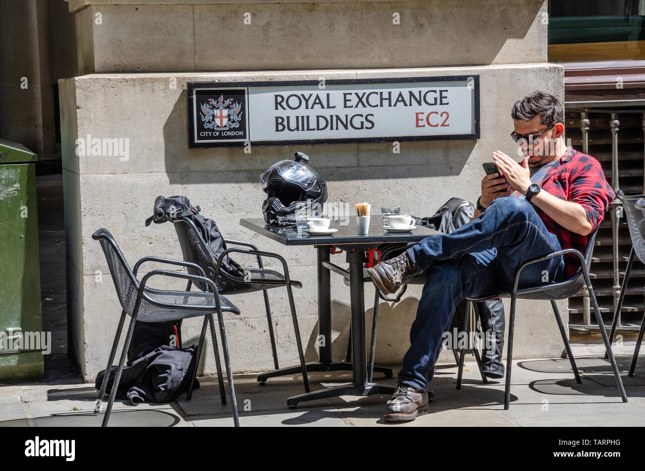 Un uomo si siede fuori ad un tavolo sulla strada al Royal Exchange edifici a Londra. Sul tavolo sono due tazze di caffè e un casco. Foto Stock