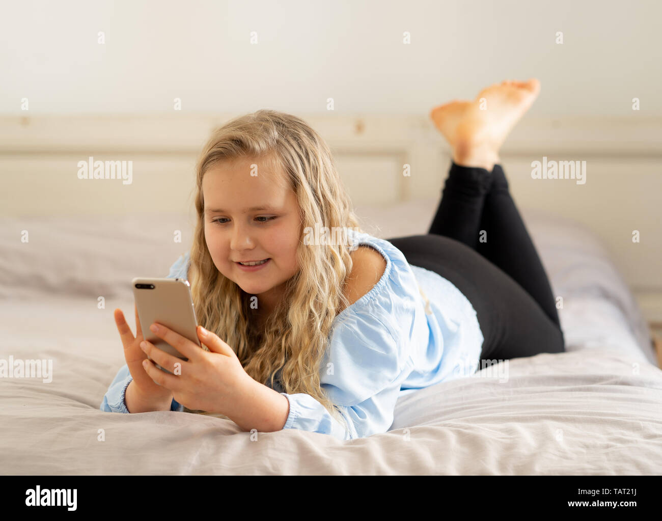 Bella bionda felice bambina gioca sul telefono cellulare. Carino il bambino in chat su internet con lo smartphone social media app giacenti nel letto di casa. Foto Stock