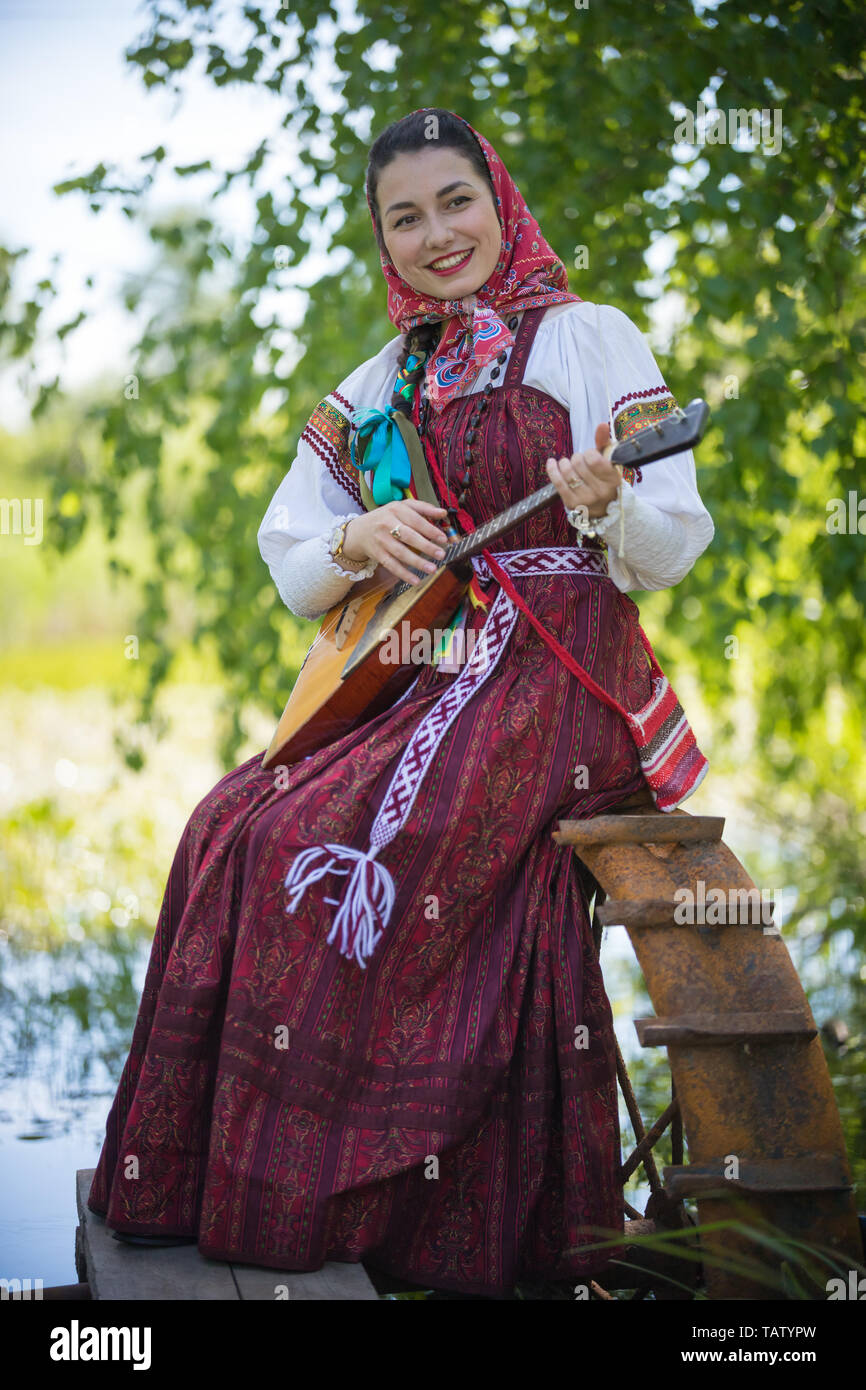Giovane donna romantica in tradizionali abiti russo si siede sul piccolo molo vicino al lago, e riprodurre musica su balalaika - colpo verticale Foto Stock