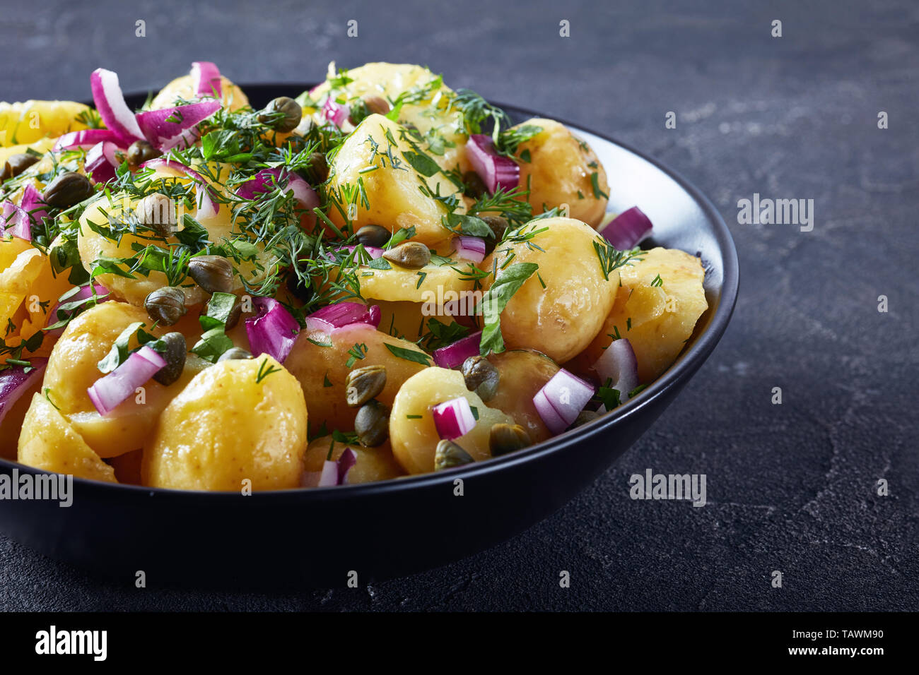 Close-up di salato tedesco nuova insalata di patate con cipolla rossa, capperi, verdi e mostarda condimento di aceto in una ciotola di nero su un tavolo in cemento, vista dal Foto Stock