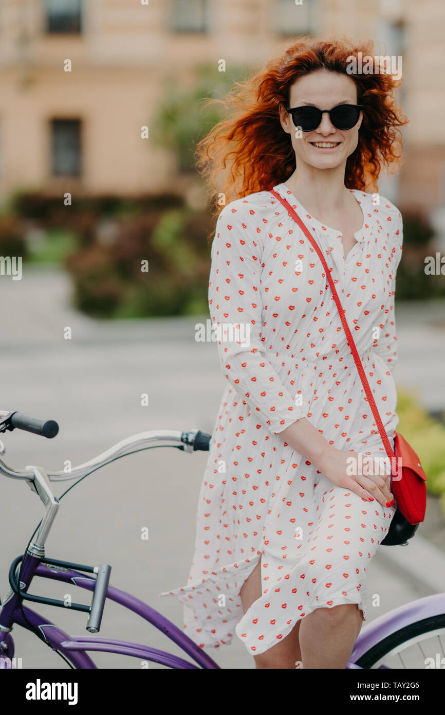 Foto di piacere dai capelli rossi donna con dolce sorriso, trascorre il tempo libero equitazione bicicletta sulle strade della città durante la soleggiata giornata di vento, indossa estate elegante DRE Foto Stock