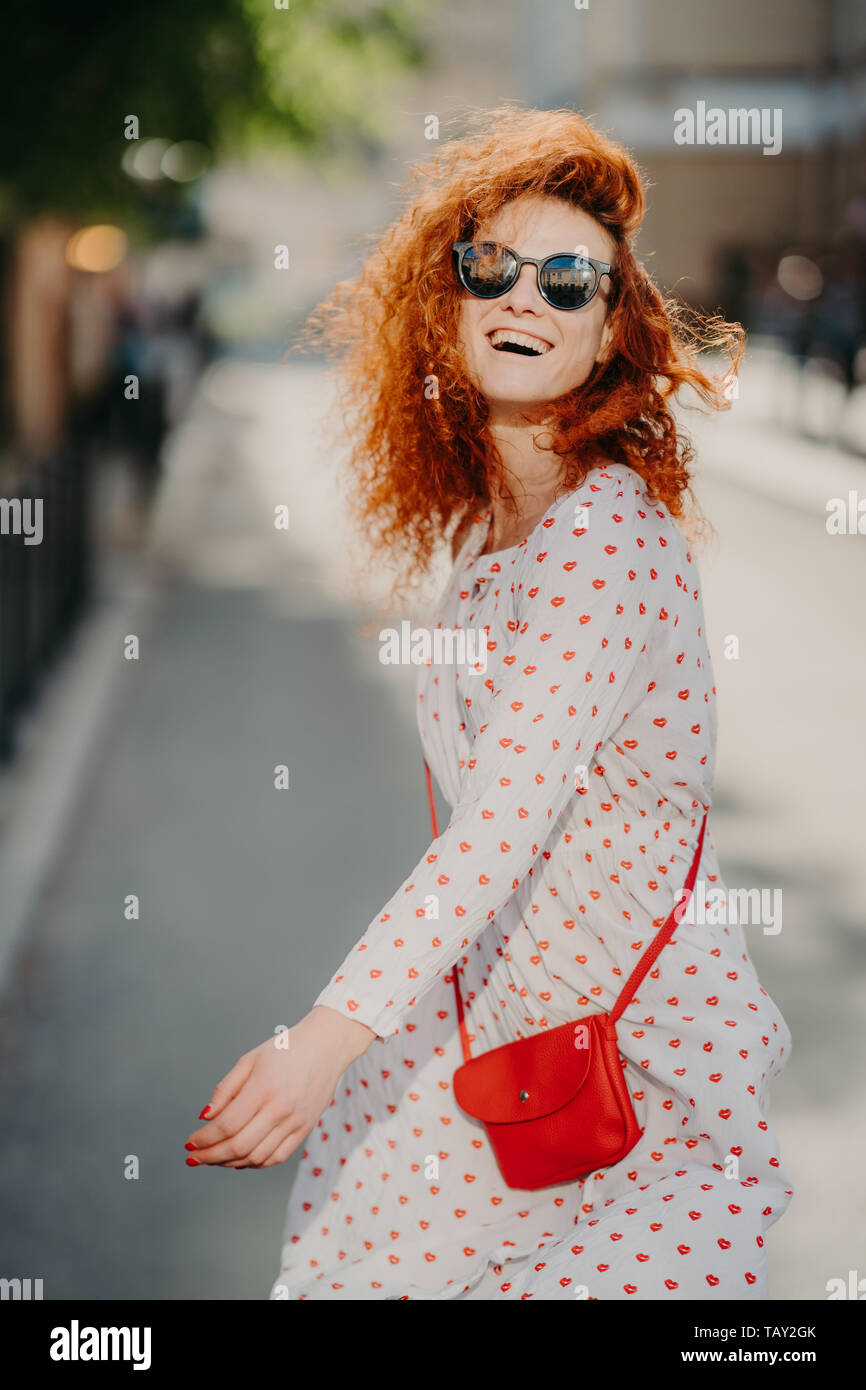 Felice di spensierata redhead donna ride positivamente, trascorre il tempo libero outdoor in città, indossa gli occhiali da sole, manica lunga, vestito porta sacchetto rosso, è divertente, godeteveli Foto Stock