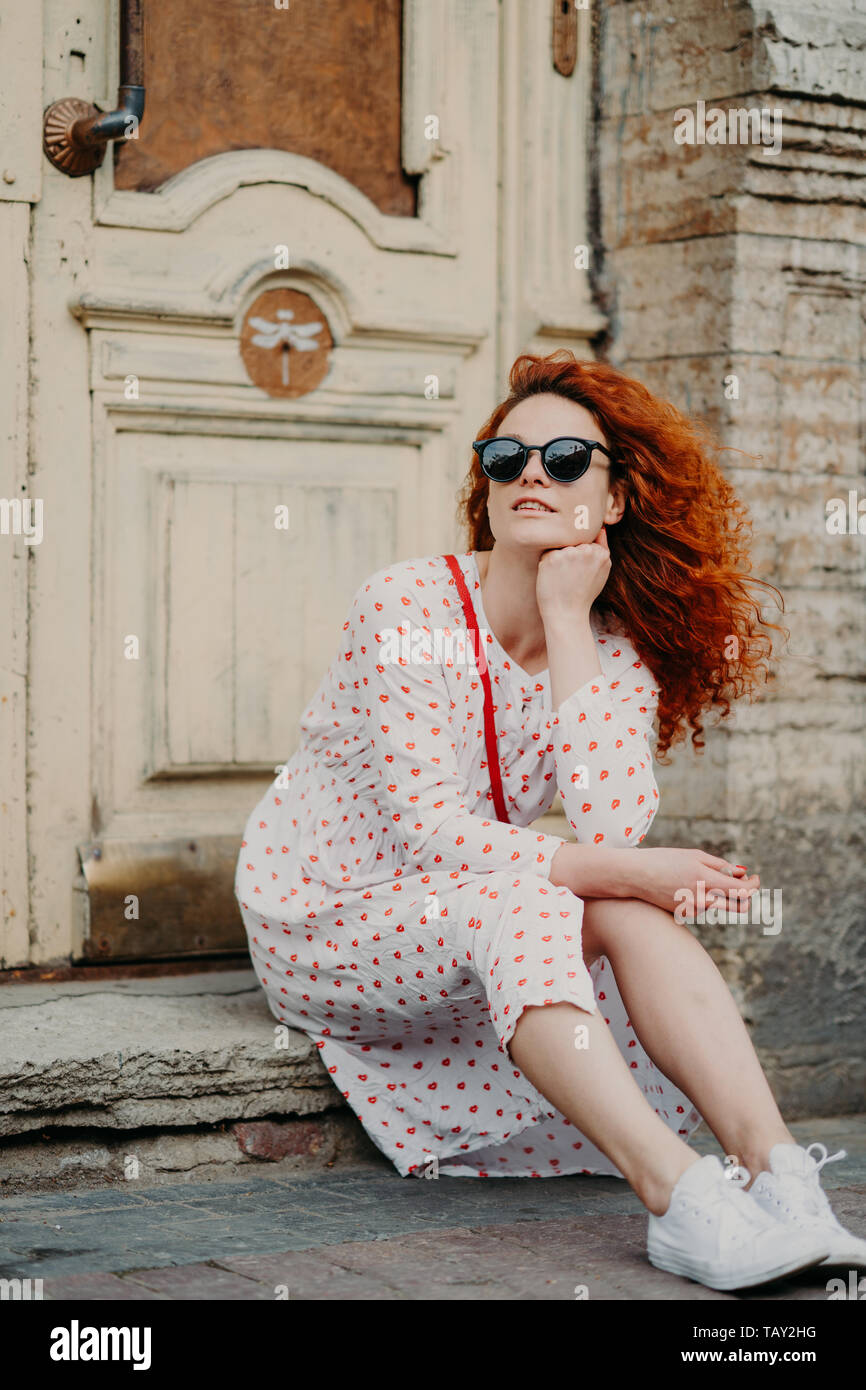 Dai capelli rossi premurosa donna ha riposo dopo le passeggiate pone vicino al vecchio edificio, siede sulla soglia, indossa gli occhiali da sole e vestito di bianco e sneakers, esplora Foto Stock