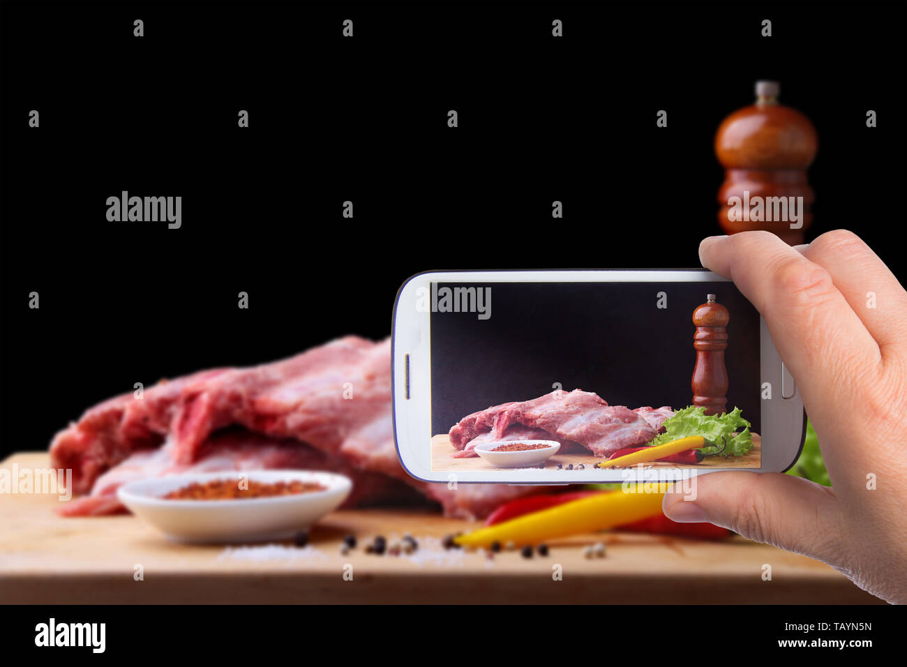 Le mani di uomo con lo smartphone tenendo foto Raw nervature su un rustico tagliere con sale e pepe e il macinino per spezie . Sfondo nero per la copia Foto Stock