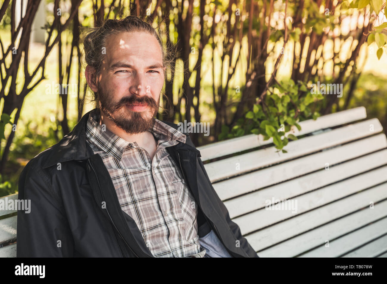 Giovane uomo barbuto si siede su un banco di bianco in estate park, ritratto all'aperto Foto Stock