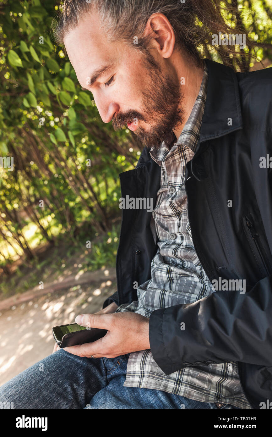 Giovani barbuto uomo sorridente con uno smartphone si trova in un parco di estate, verticale ritratto all'aperto Foto Stock