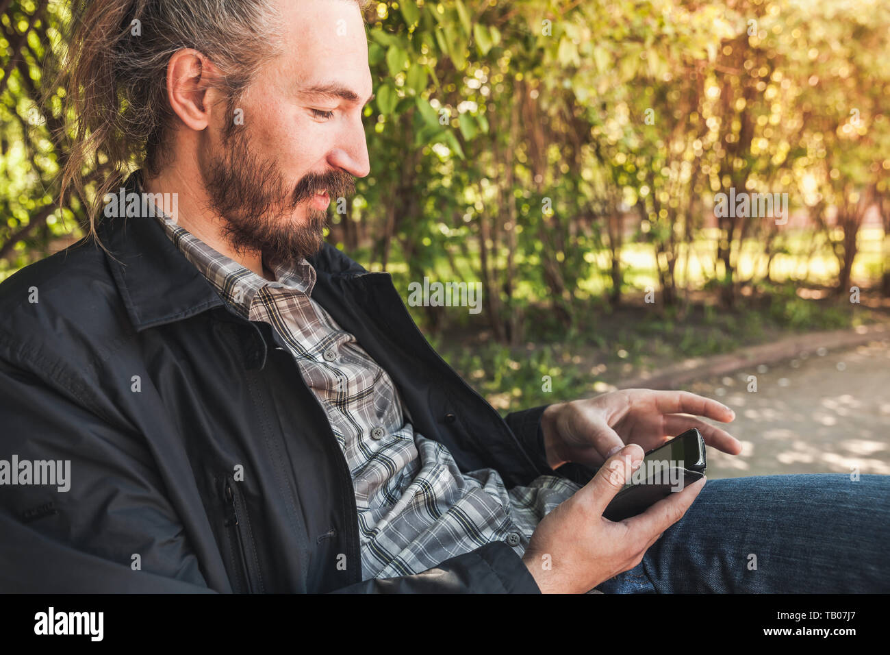 I giovani adulti barbuto uomo sorridente con uno smartphone si trova in un parco di estate, verticale ritratto all'aperto Foto Stock
