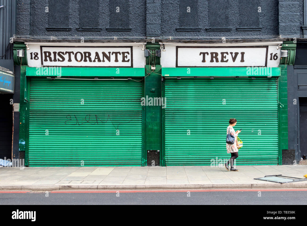 Una donna che cammina passato Ristorante Trevi, un ristorante italiano a Highbury, angolo chiuso con il suo verde-metallo verniciato serrande giù, London, Regno Unito Foto Stock