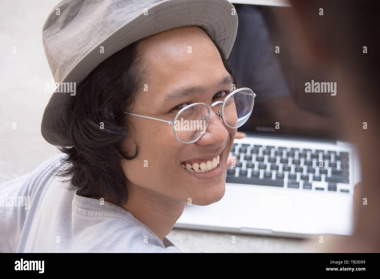 Giovane uomo asiatico con cappello e occhiali sorridendo a un amico con il computer portatile in background Foto Stock