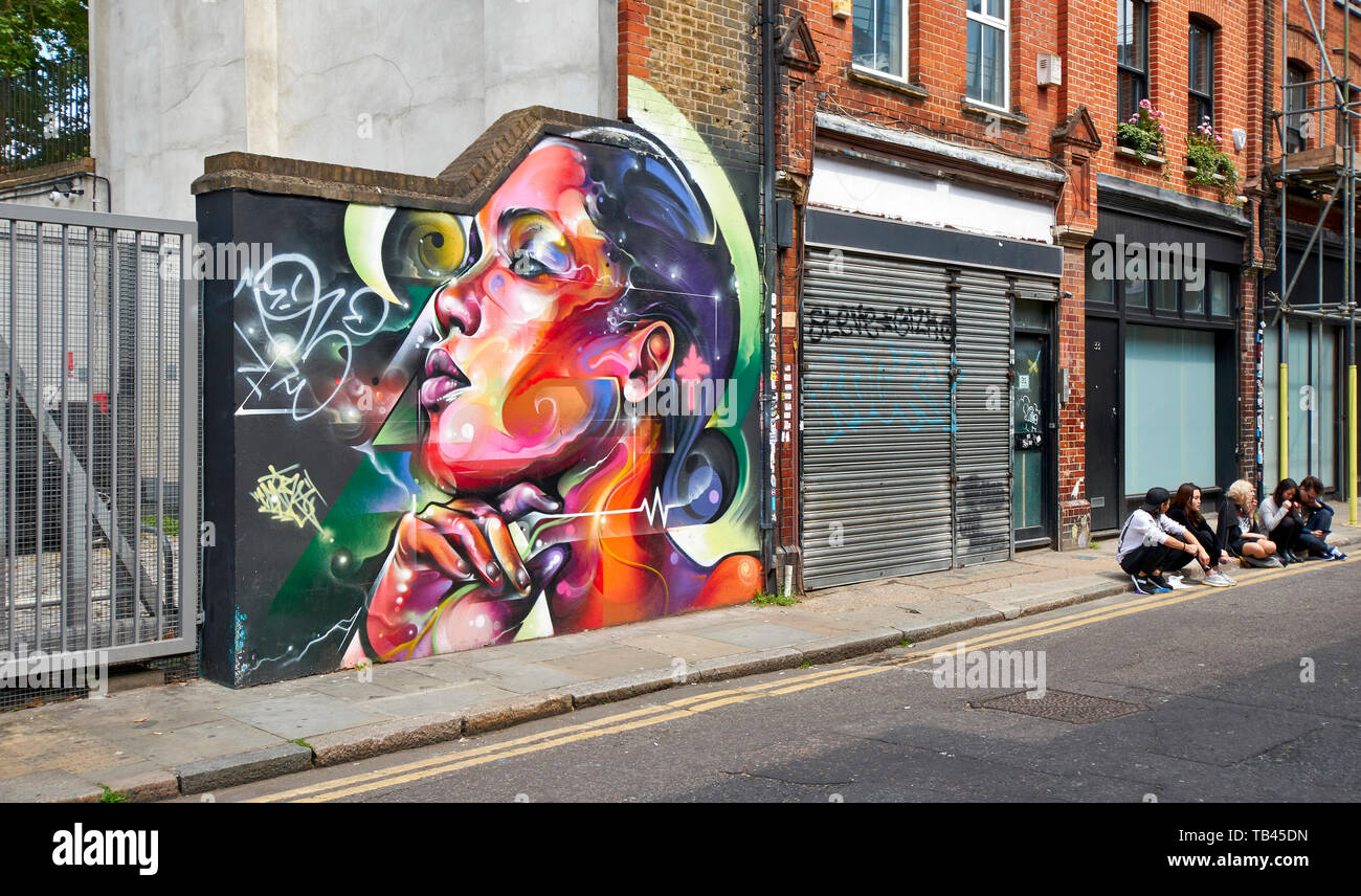Londra SPITALFIELDS Brick Lane Street con muro di arte e cinque persone sedute sul pavimento Foto Stock
