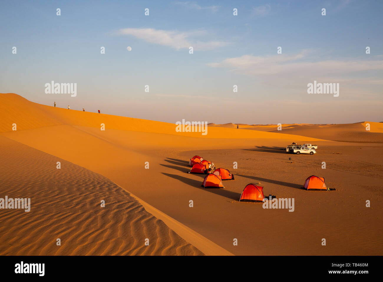Le tende si accamparono per campeggio selvaggio in serata sole nel deserto occidentale del Sudan. Quattro figure salire la cresta di una duna di sabbia come la luna tramonta Foto Stock