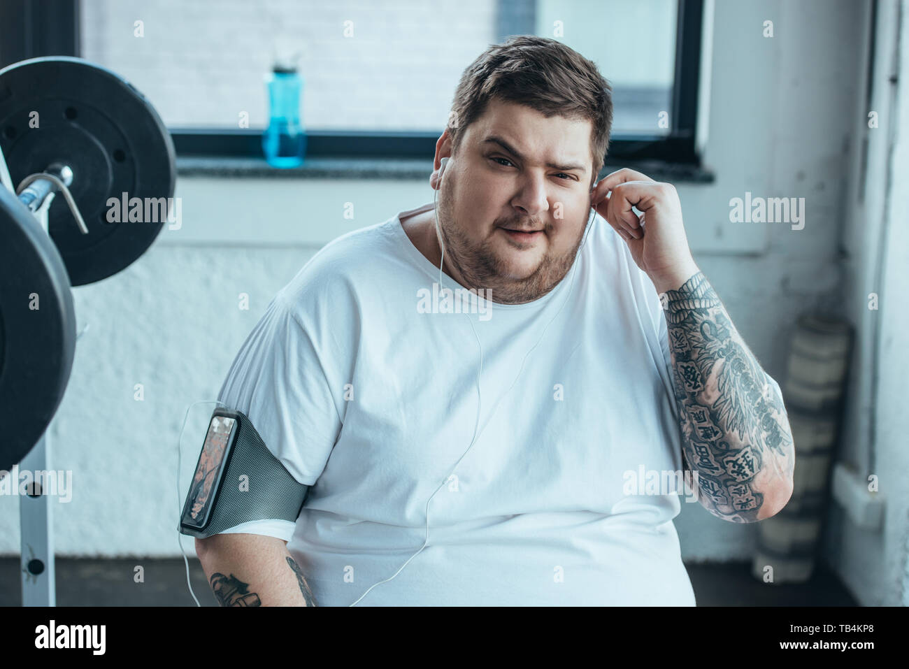Il sovrappeso uomo nella fascia da braccio per smartphone guardando la fotocamera pur mettendo su auricolari in corrispondenza della palestra Foto Stock