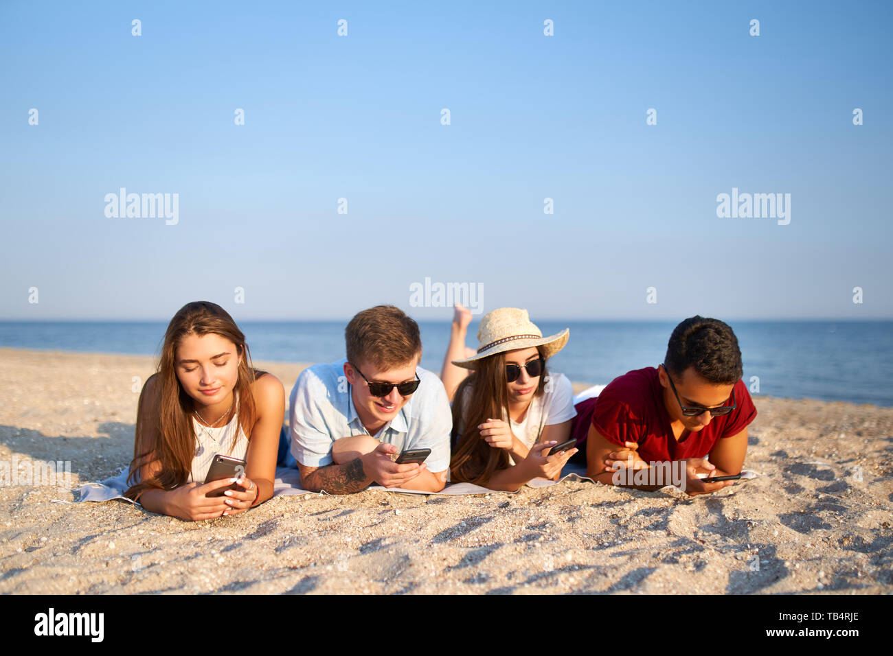 Gruppo di millenials smartphone utilizzando la posa insieme sul telo da spiaggia vicino al mare in estate il tramonto. I giovani tossicodipendenti da mobile smart phone. Sempre Foto Stock