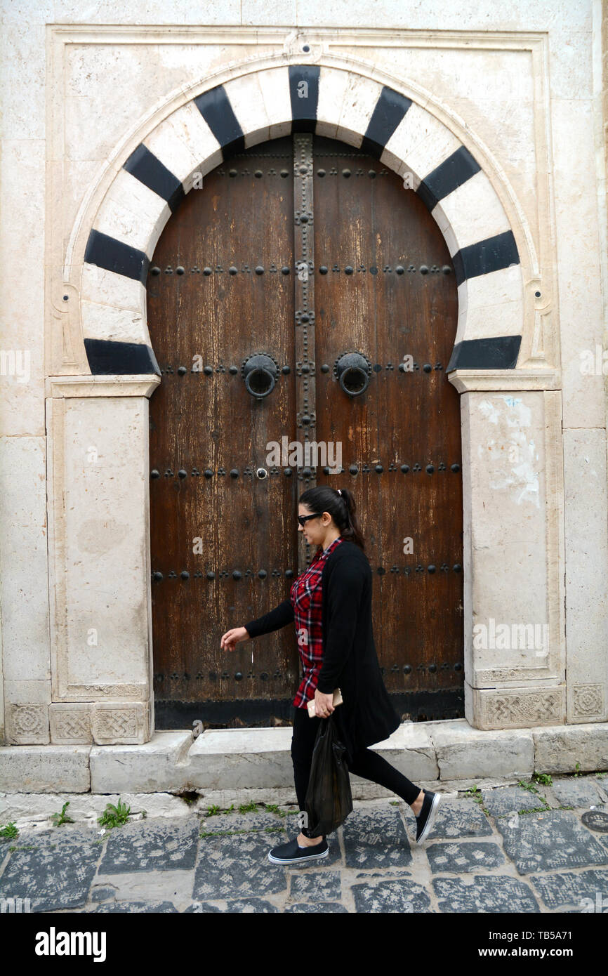 Un tunisino donna cammina oltre la tradizionale in legno chiodati porta di una casa del XVI secolo in un vicolo della medina (città vecchia) di Tunisi, Tunisia. Foto Stock