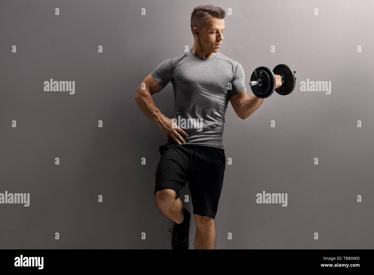 Giovane uomo muscolare di sollevare un manubrio contro un muro grigio Foto Stock