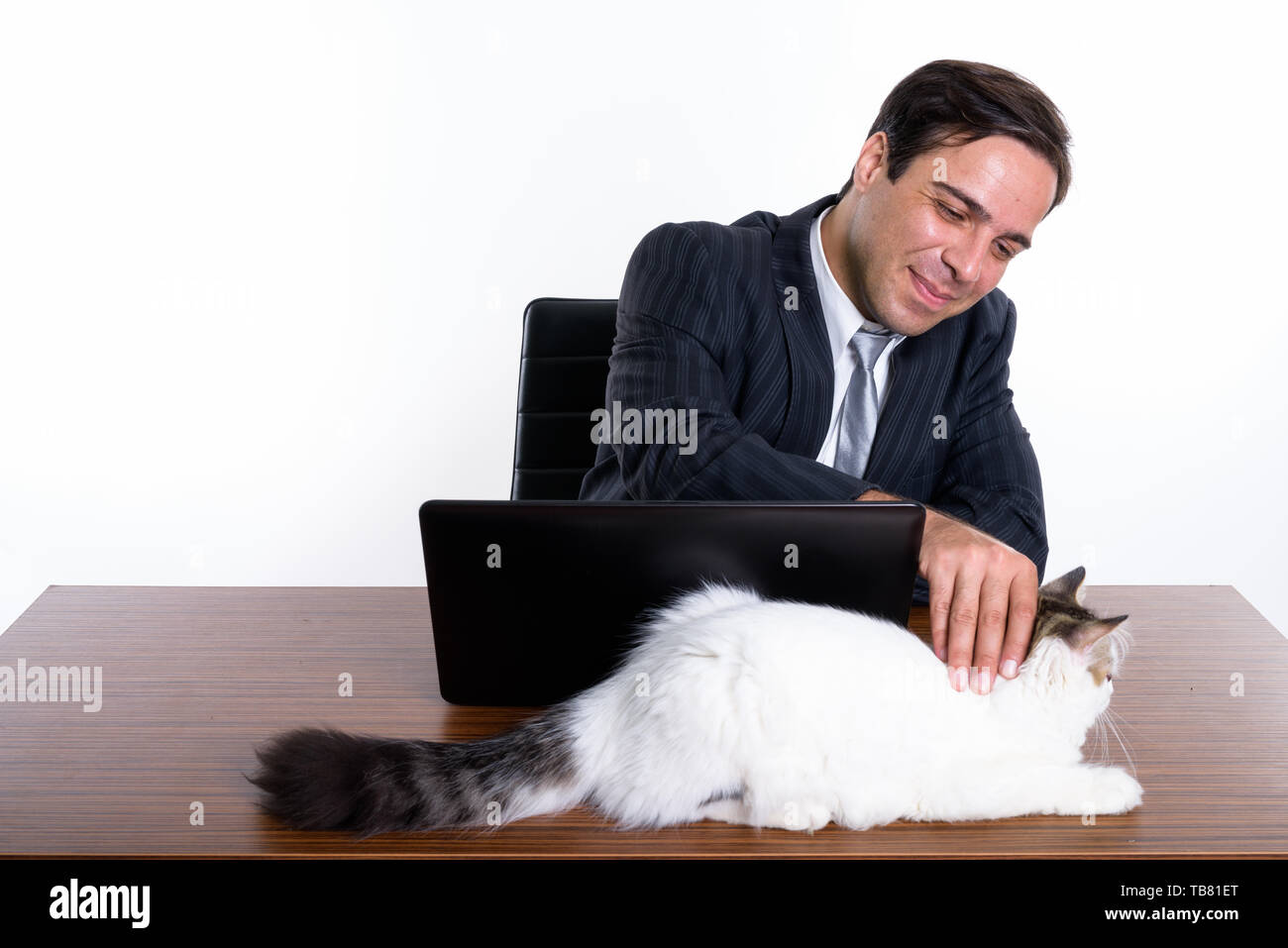 Studio shot del giovane imprenditore persiano toccando cute cat mentre è seduto con il portatile sul tavolo di legno contro uno sfondo bianco Foto Stock