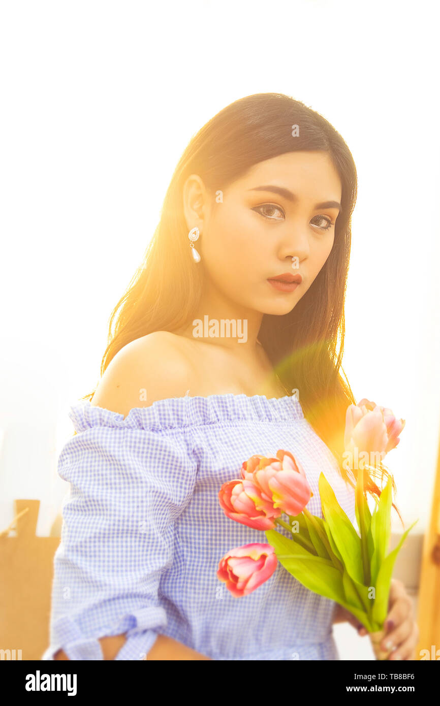 Belle Donne asiatiche in vestito blu con red tulip fiori in mani con la luce del sole di mattina da finestra Foto Stock
