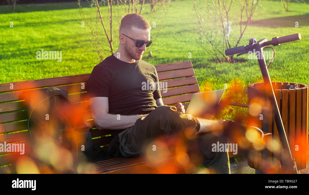 Giovane uomo attraente con laptop e moderna kick elettrico scooter seduti al parco contemporaneo al banco di lavoro e il freelance o business man workin Foto Stock