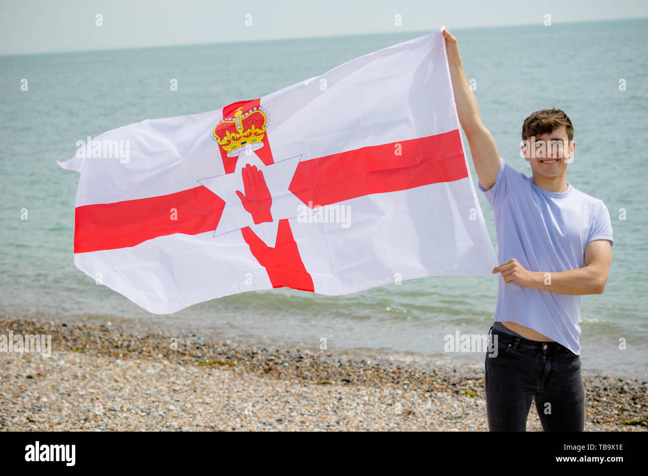 Giovane adulto maschio caucasico tenendo premuto su una spiaggia tiene la bandiera dell'Irlanda del Nord Foto Stock