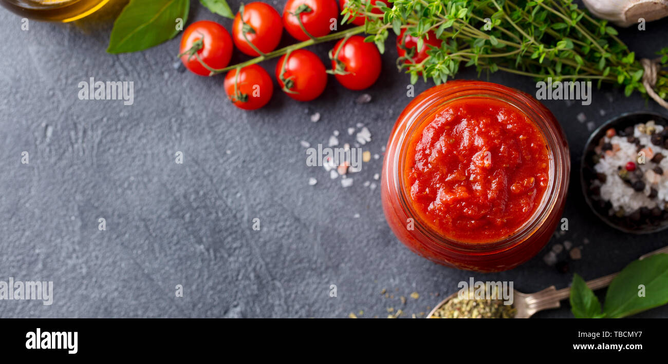 Tradizionale salsa di pomodoro in un vasetto di vetro con erbe aromatiche, pomodori e olio di oliva. Vista dall'alto. Copia dello spazio. Sfondo di ardesia. Foto Stock