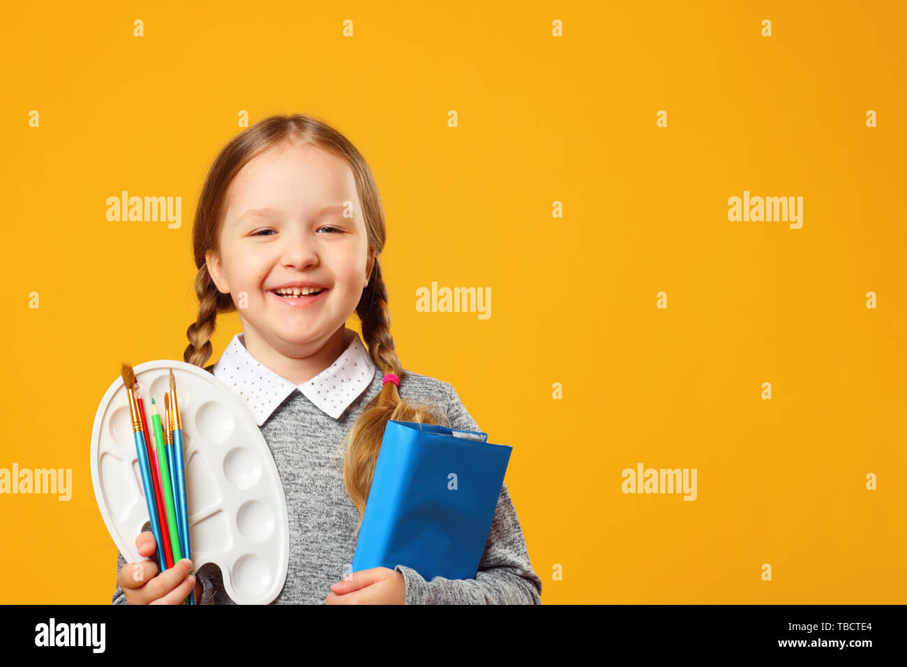 Ritratto di un allegro bambina su uno sfondo giallo. Schoolgirl tiene un libro, matite, pennelli e una tavolozza. Il concetto di istruzione. Copia spa Foto Stock